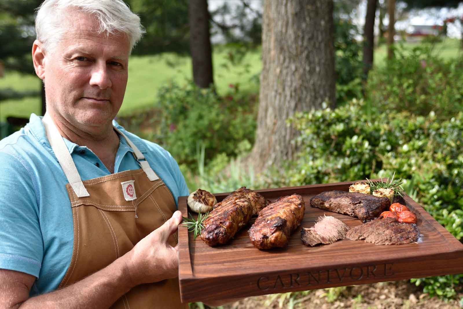 A man holding a meat board with steak, chicken and vegetables on it. The scene captures the warmth of home cooking, showcasing the importance of personalized kitchen utensils in creating memorable meals for loved ones.