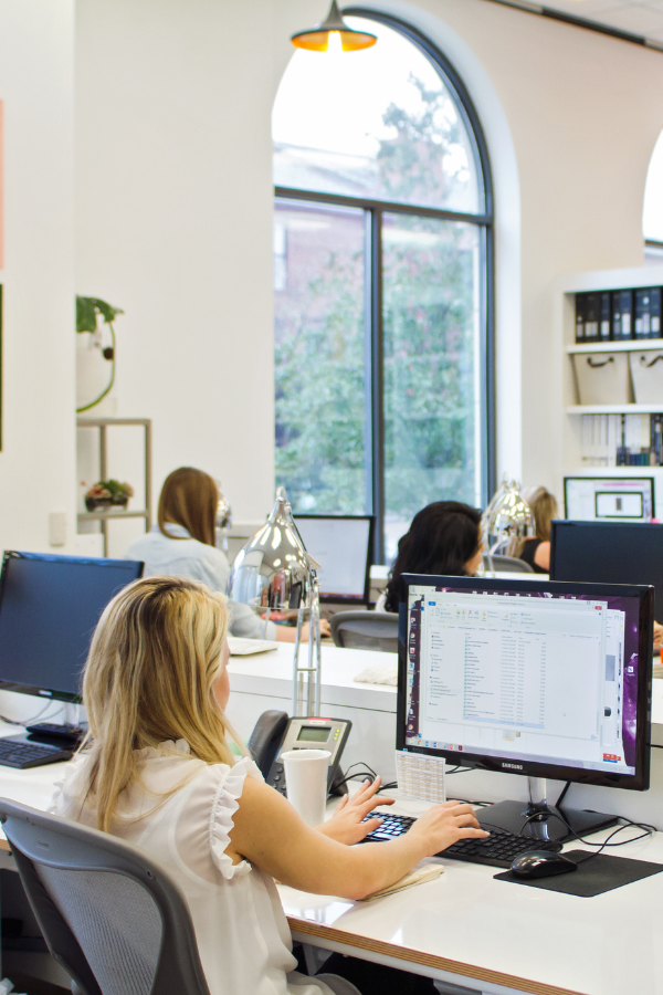 Designers work at desks in a bright, modern office with large arched windows, desktop computers, and task lighting.