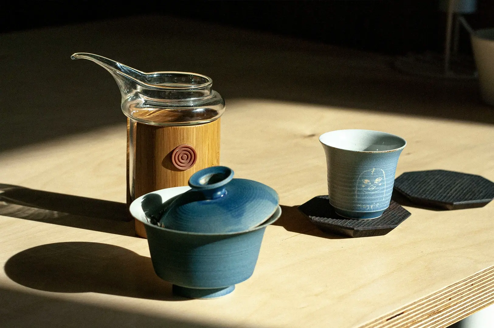 A close-up, high-angle shot of a minimalist tea setup on a light wooden surface. A clear glass teapot with a bamboo sleeve sits next to a small blue lidded ceramic cup (gaiwan). To the right, a matching blue cup featuring a whimsical line-art drawing of a face and the word "Magpie" rests on a black, hexagonal coaster. Natural sunlight casts sharp, dramatic shadows across the scene.
