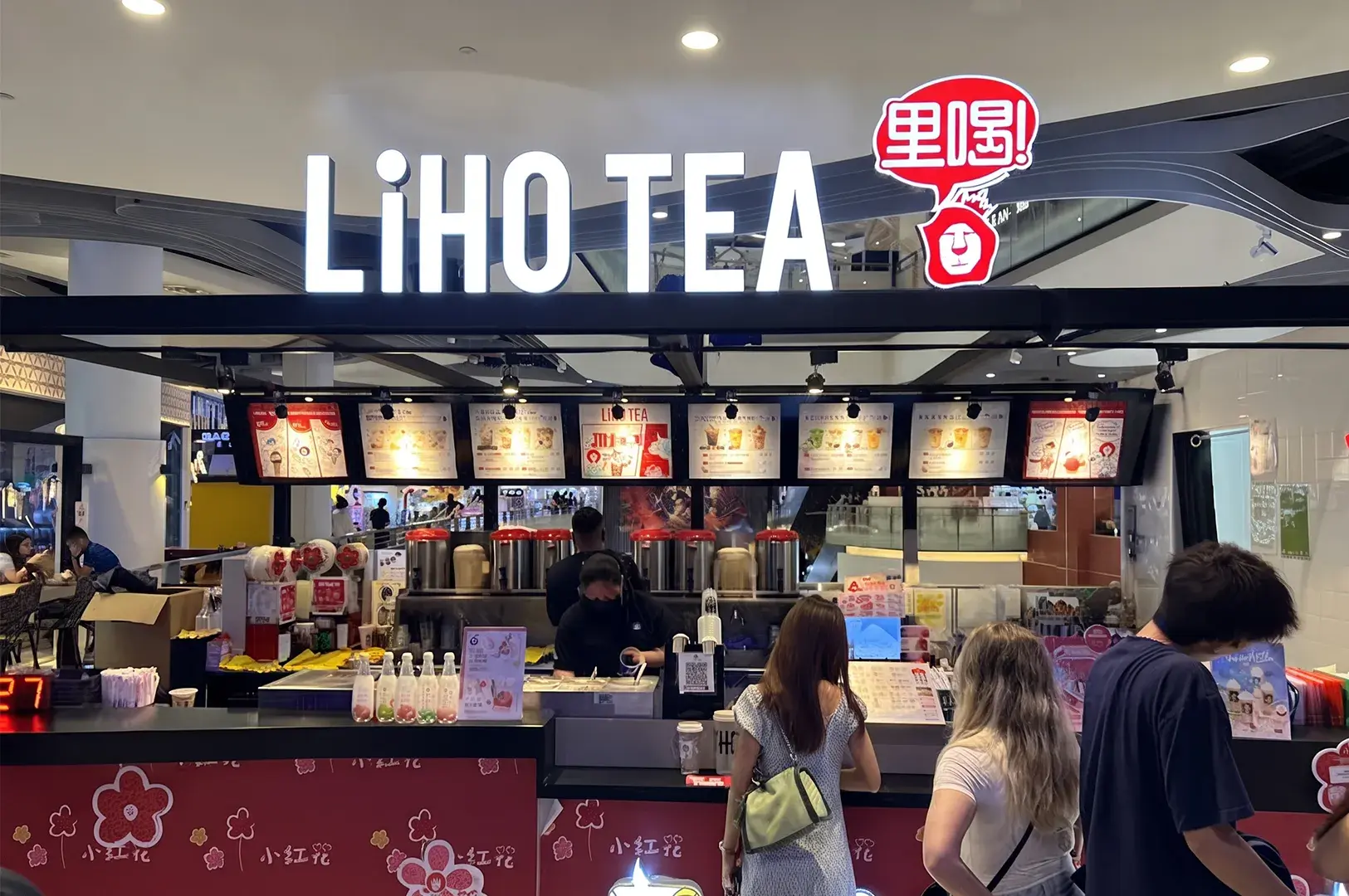 Short Alt Text (Best for general web/social media) Storefront of a LiHO TEA kiosk in a shopping mall, featuring bright white illuminated signage, red brand logo, illuminated menu boards, and customers lining up at the counter.
