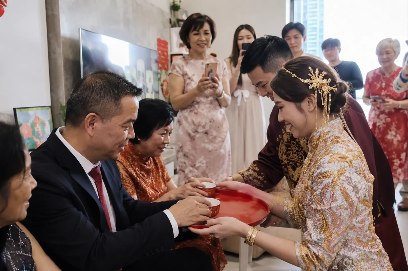 In a brightly lit room, a bride and groom present a red tray holding two small tea cups to an older couple sitting before them. The bride, viewed in profile from the right, smiles while wearing a heavily embroidered gold, white, and pink traditional Qun Kwa dress with an ornate gold hairpin structure in her hair. The groom, partially visible behind her, wears a dark burgundy tunic with gold dragon embroidery. They are serving tea to a man in a black suit with a red tie and a woman in an orange patterned dress. In the background, family members smile and capture the moment with their smartphones.