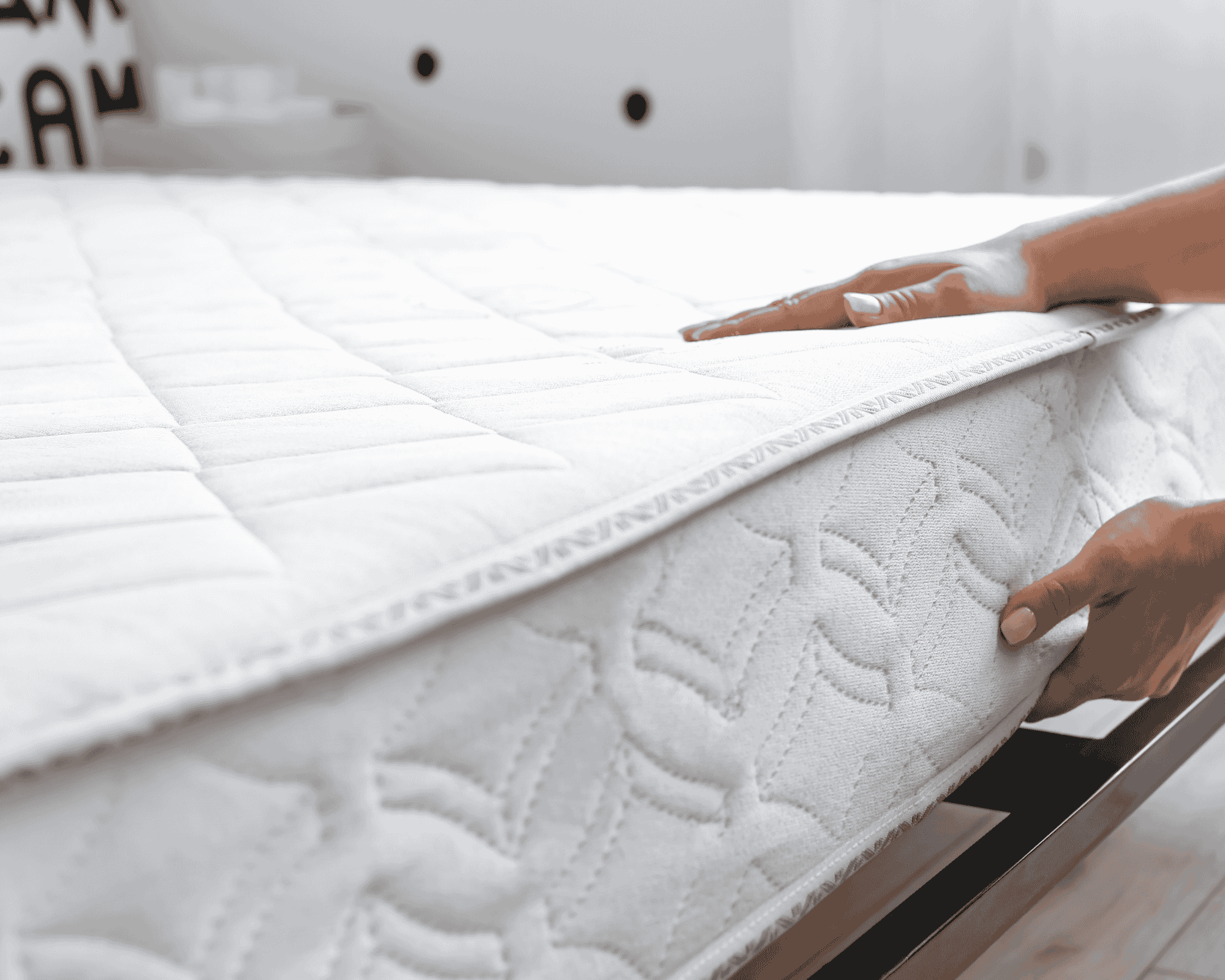Close-up of a person adjusting or placing a clean, white mattress on a bed frame with both hands. The mattress has a textured, quilted surface in a bright, well-lit bedroom