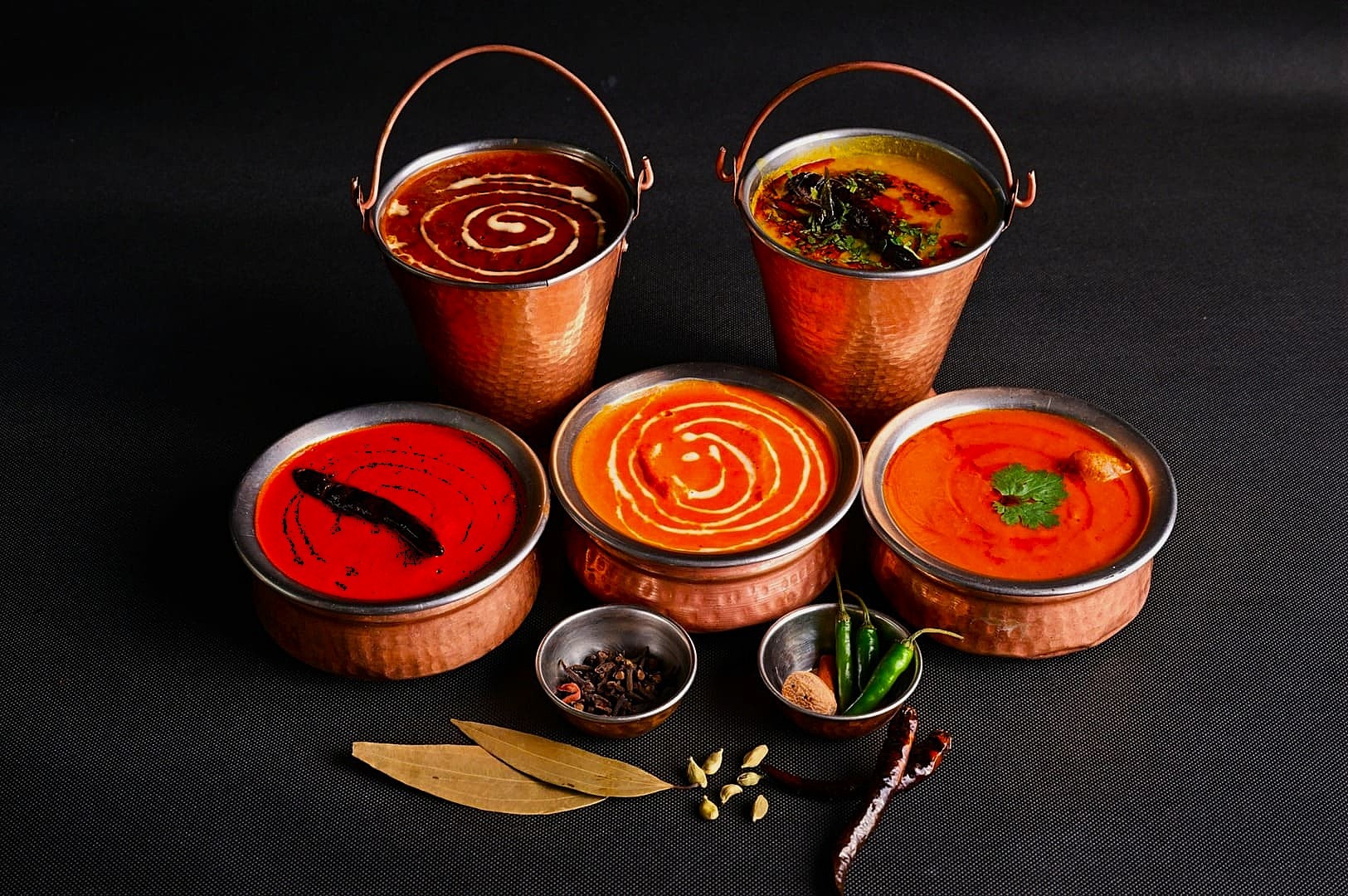 Five copper bowls and buckets filled with colorful Indian curries are arranged against a dark background, with spices and chilies placed in front.