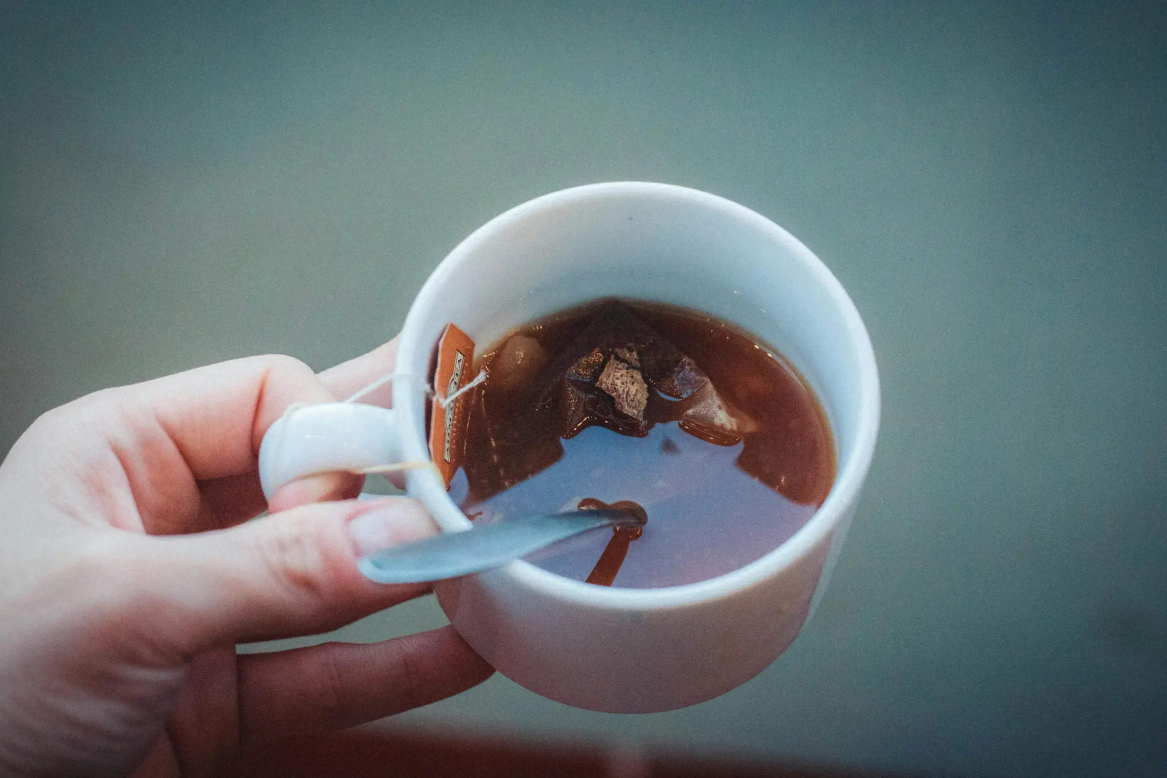 A hand holds a white mug filled with dark amber tea, with a submerged tea bag and a small metal spoon resting inside. The scene is shot from a top-down perspective against a soft, muted blue-grey background.