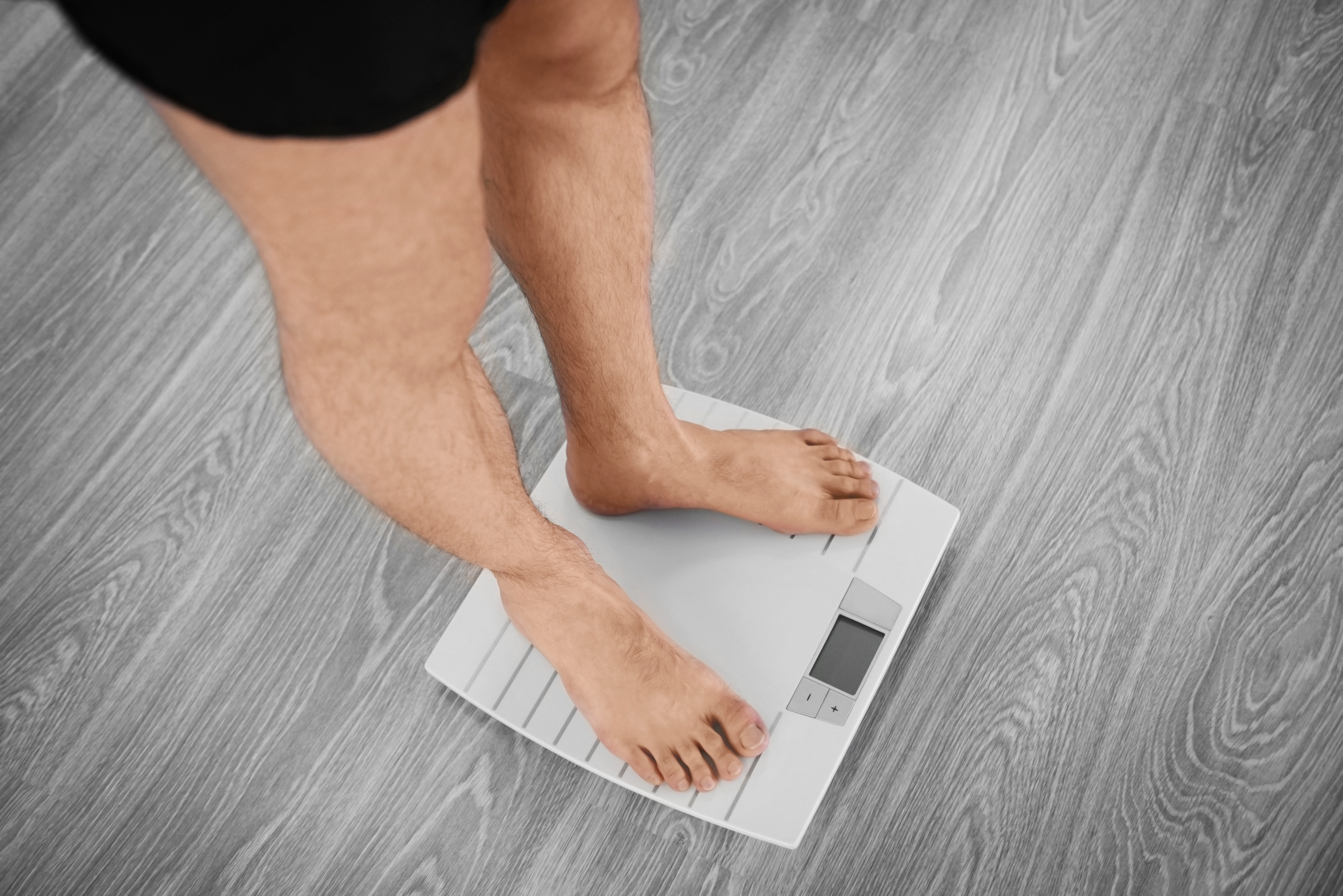 Lower body shot of a white man with slim legs, standing on an electronic weight scale which has been placed on a grey, artificial wood floor