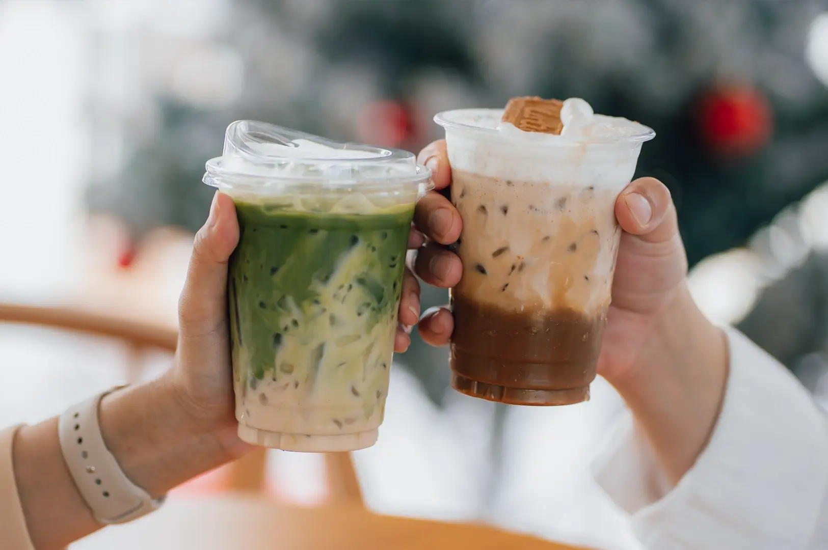 Close-up eye-level shot of two hands holding iced matcha latte and iced caramel coffee in clear takeaway cups inside a café, with a softly blurred background.