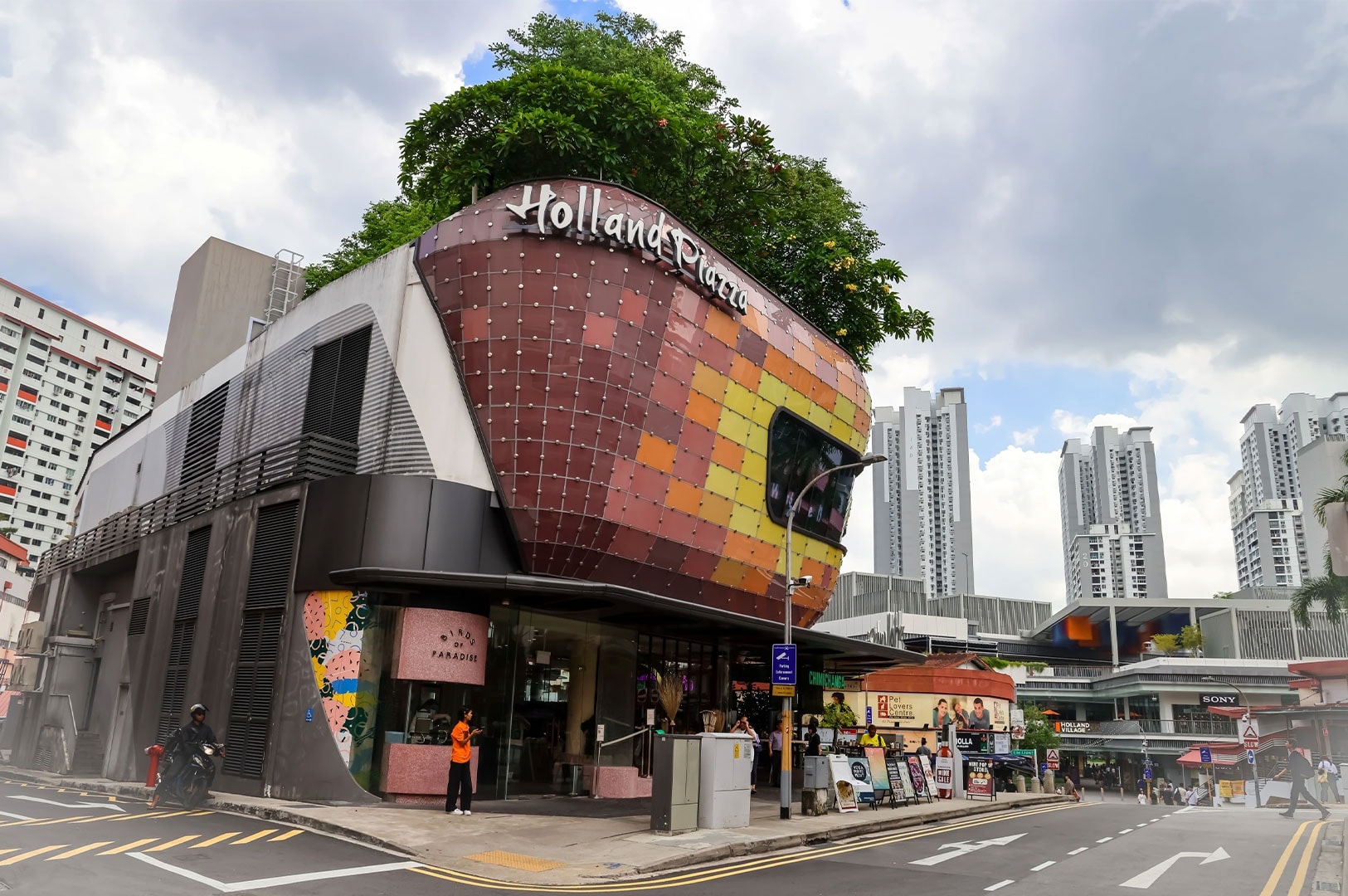 Modern, distinctive Holland Village Plaza building with a colorful, tiled facade and lush rooftop garden, set against high-rise residential buildings.