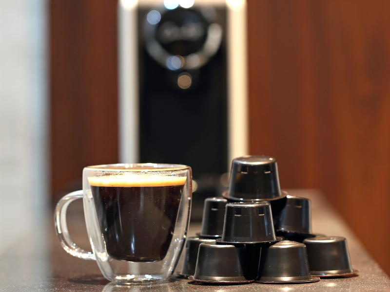 Cápsulas e xícara de café em primeiro plano e cafeteira ao fundo. Foto: grass-lifeisgood - Canva