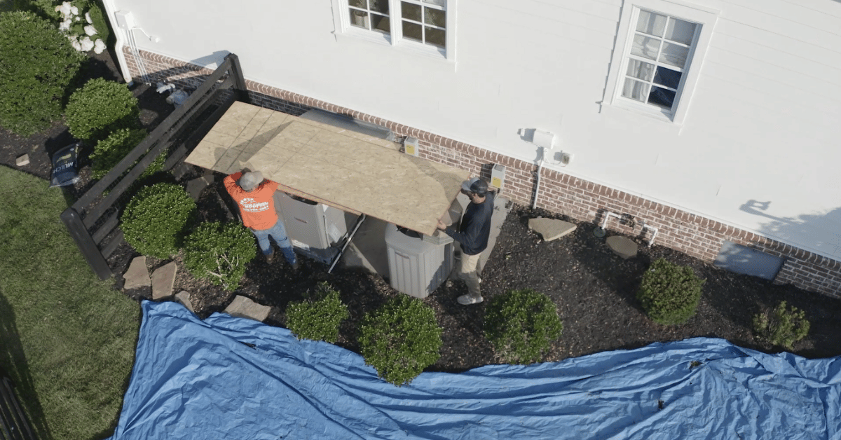 Roofers placing a large plywood board over HVAC units and utility boxes to protect equipment from falling debris during roof tear-off.