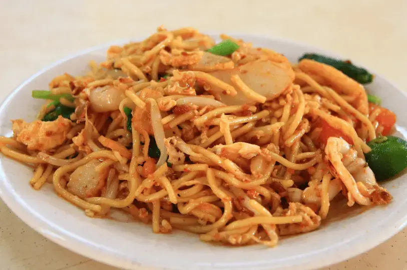 A close-up shot of a plate of Fried Hokkien Mee. The thick rice vermicelli is tossed in a savory, spiced paste and mixed with bean sprouts, prawns, fish cake slices, and garnished with fresh green vegetables.