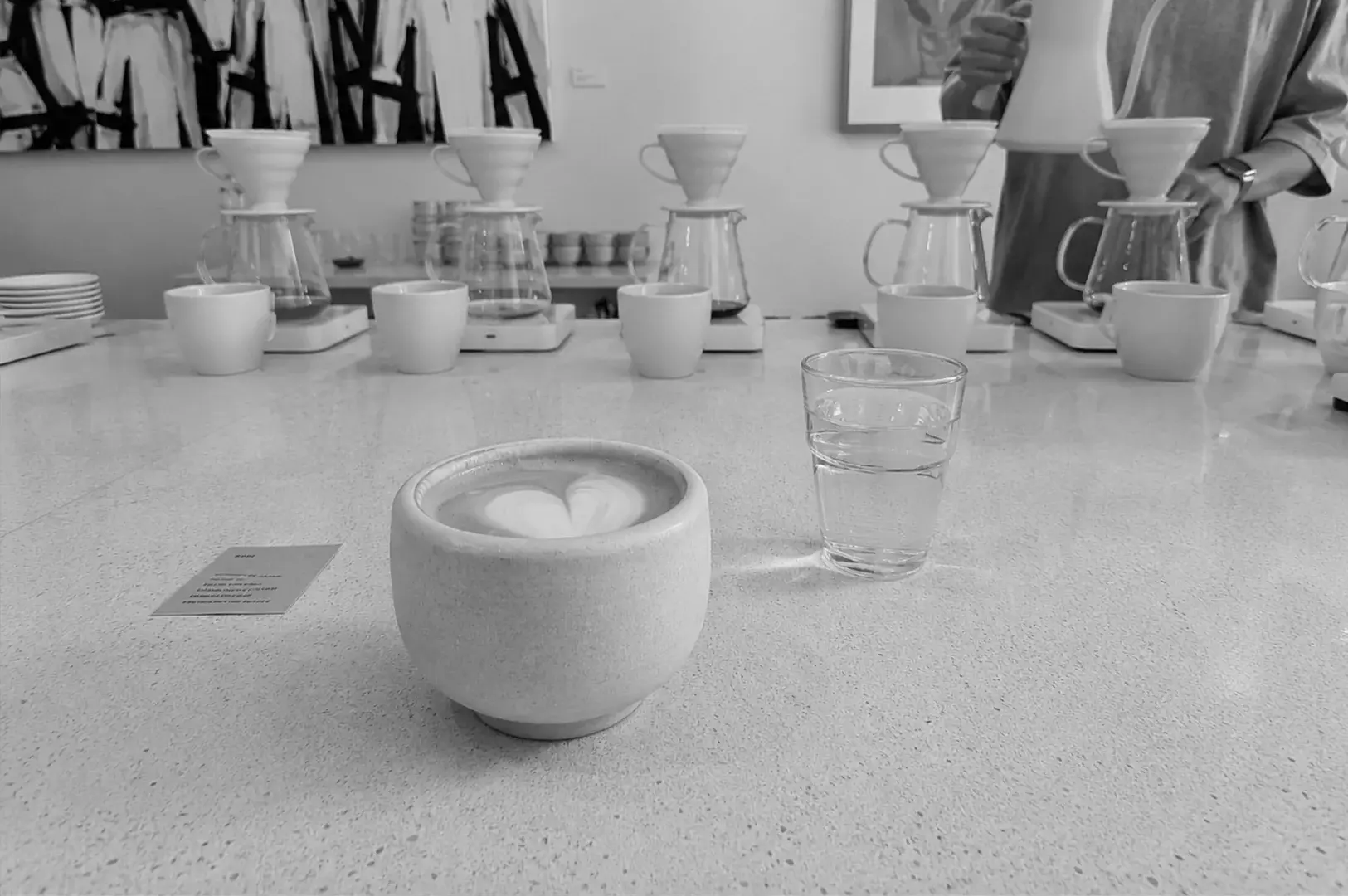 Minimalist latte with heart art placed on a bright café counter with pour-over coffee setup in the background.