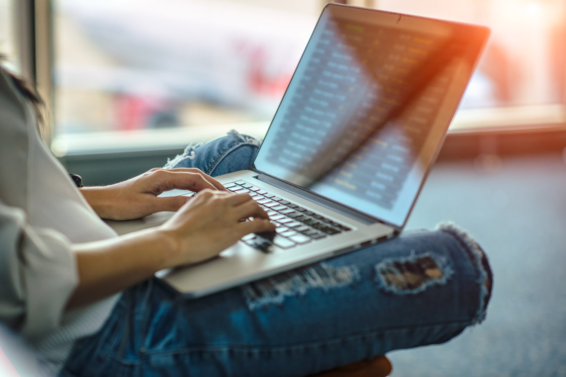 Casual woman using laptop at airport lounge, comparing broker listings and sales data