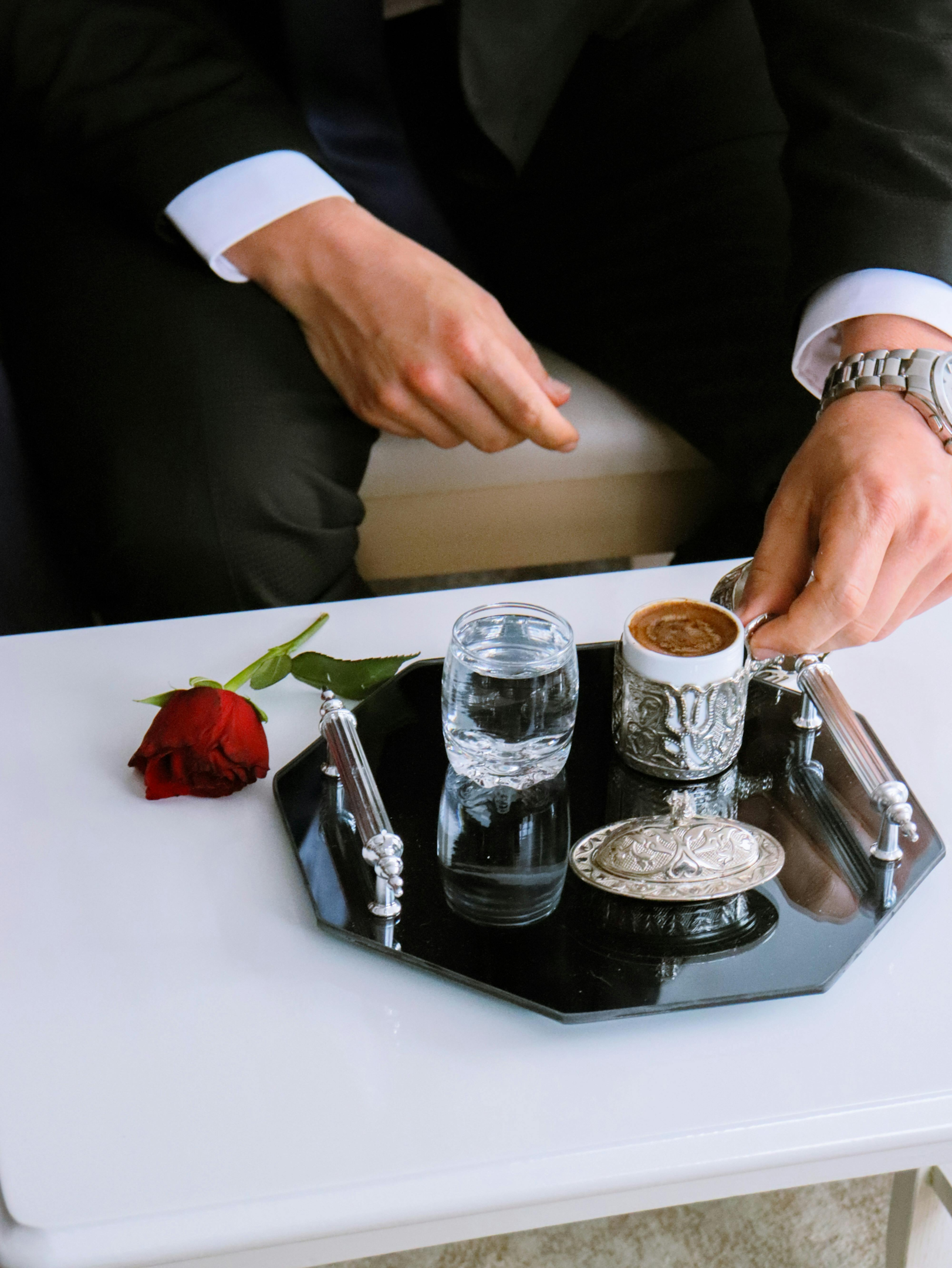 gentleman with a coffee in a fancy cup and a glass of water on elegant black tray beside a red rose