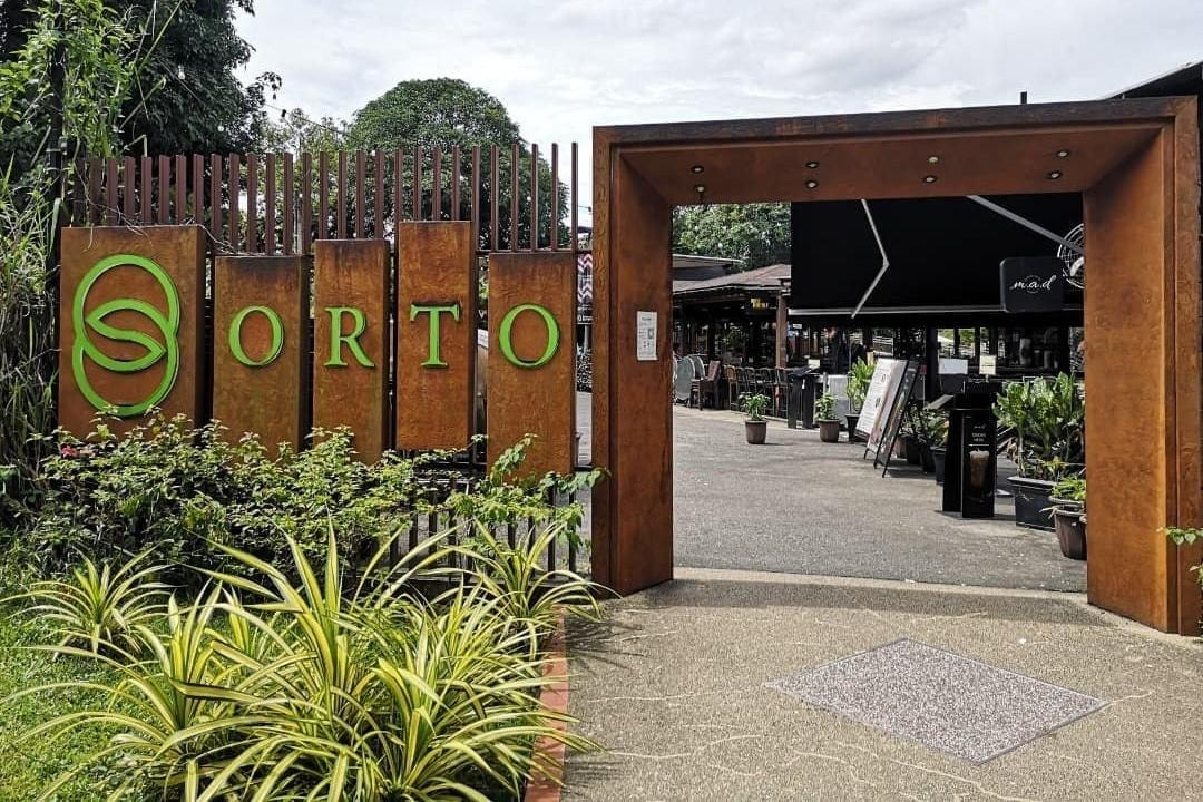  An industrial-style entrance features rusted metal panels and a large rectangular gateway that leads into an outdoor lifestyle destination. The signage prominently displays the "ORTO" logo in bright green lettering against the weathered steel background, flanked by lush tropical plants.