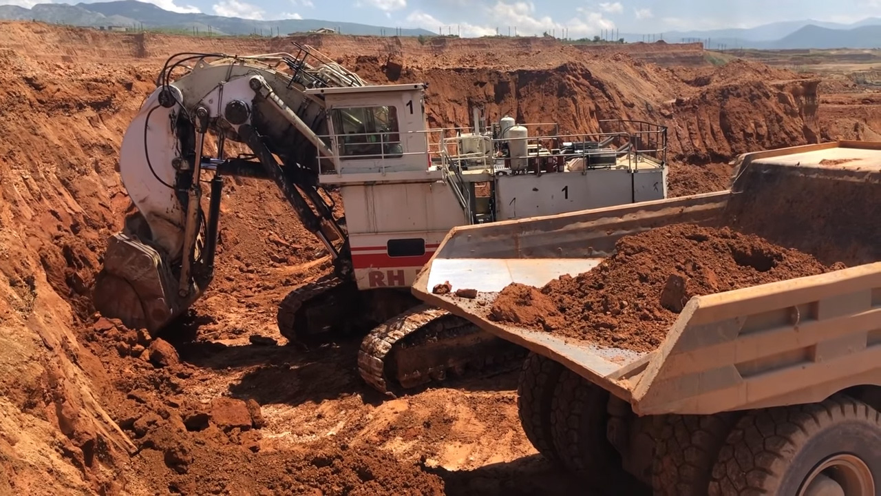 A large excavator with a bucket full of dirt is positioned over a dump truck, ready to unload the soil into the truck's bed at a construction site with red dirt and a clear blue sky in the background