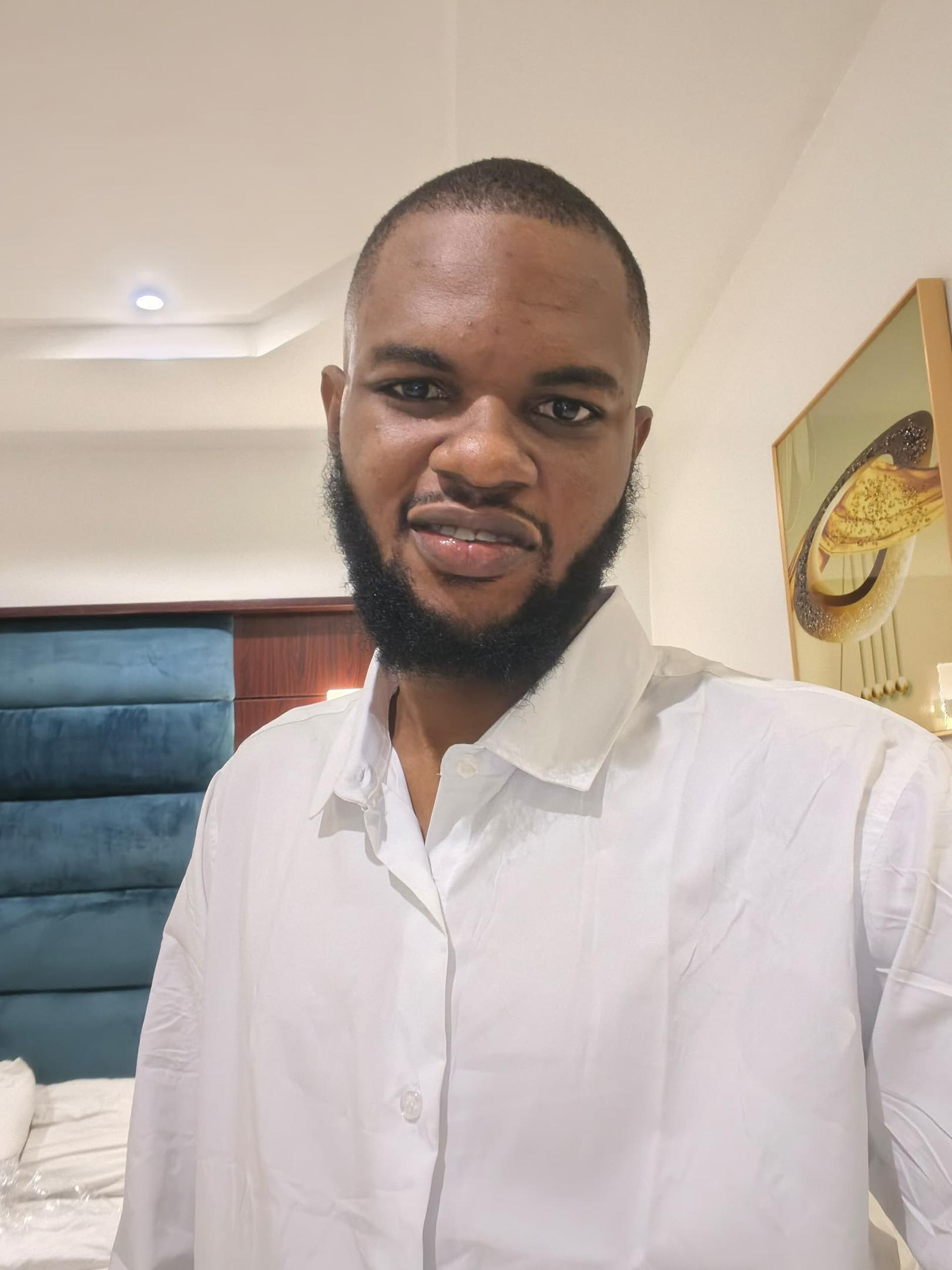 A young man with a full black beard wearing a crisp white button-down shirt, looking directly at the camera with a calm, self-assured expression, photographed in a well-appointed hotel room with a teal upholstered headboard and decorative wall art in the background.