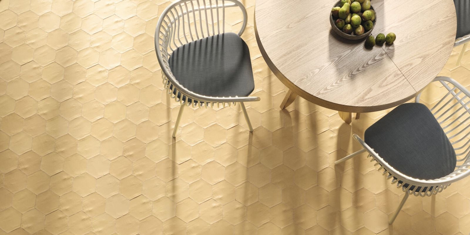Overhead view of a modern dining space featuring pale yellow hexagonal floor tiles, a round light wood table, and white rattan chairs with dark cushions.