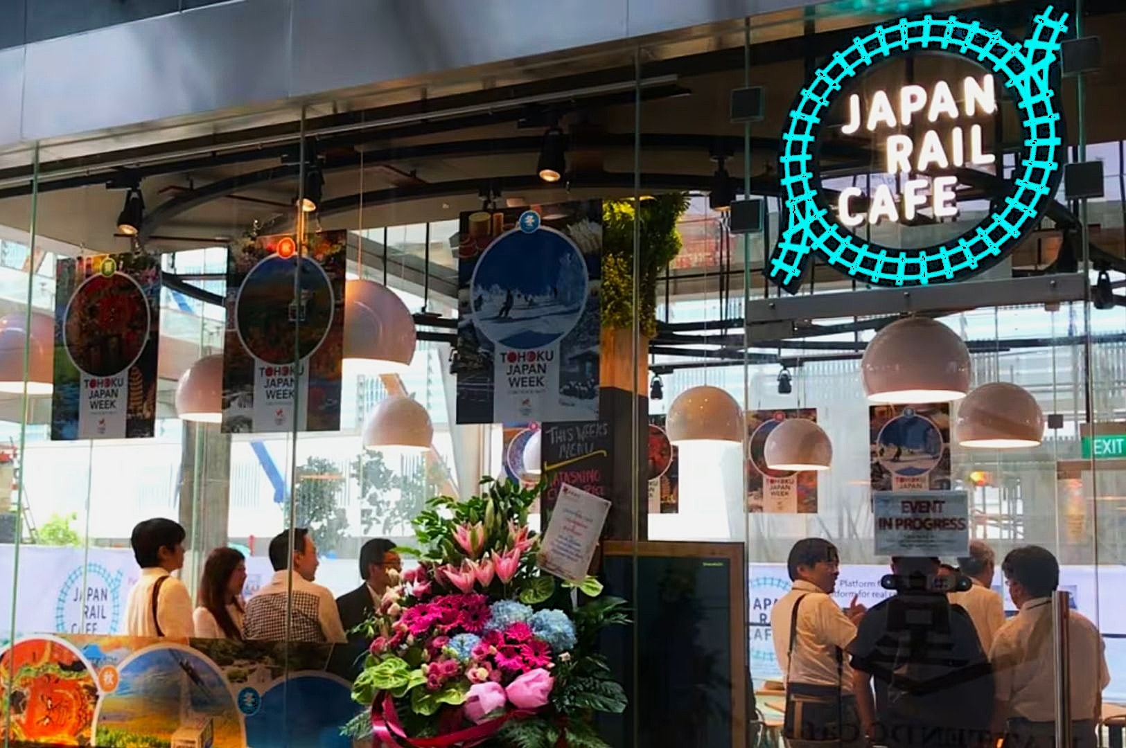 Interior shot of a Japan Rail Cafe with neon sign, colorful flower arrangement, posters for Tohoku Japan Week, and people gathering inside.