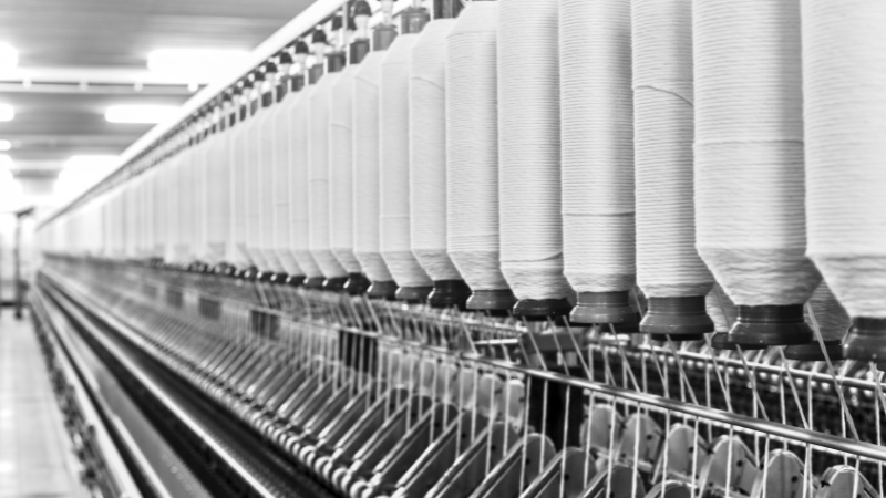 A grayscale image shows a long row of large white spools of thread on a spinning machine in a textile factory.