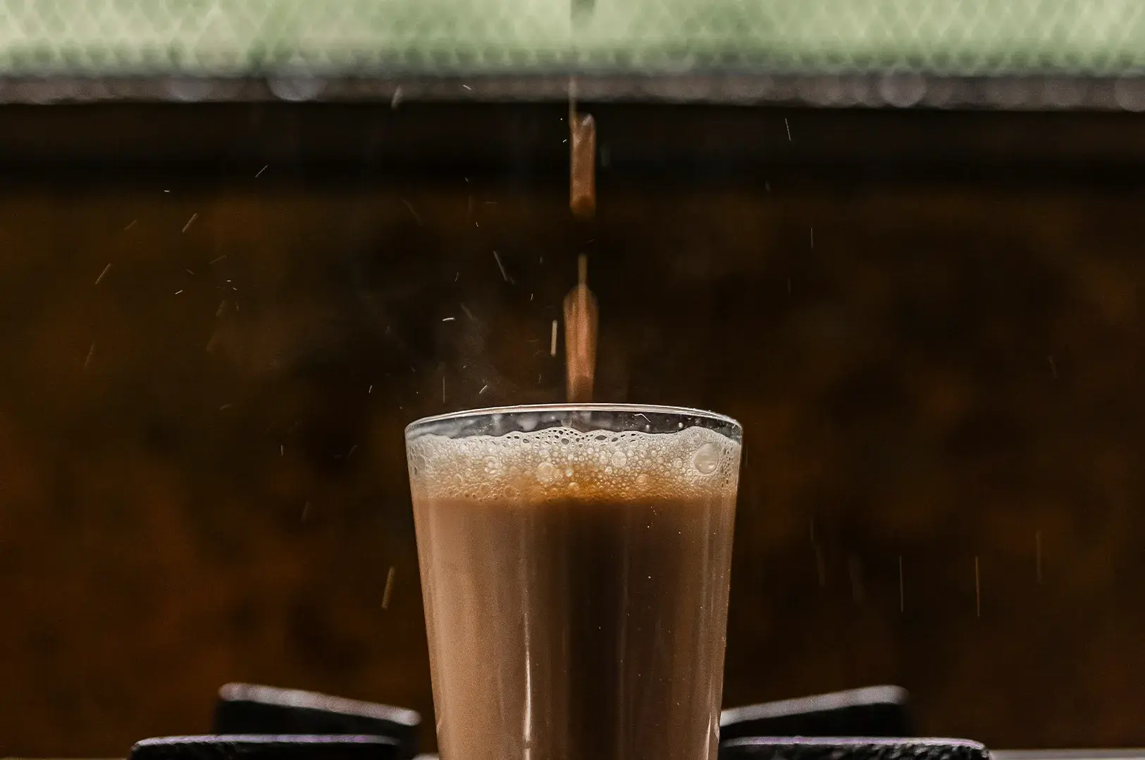 Action shot of hot tea being poured from above into a clear glass filled with frothy chai.