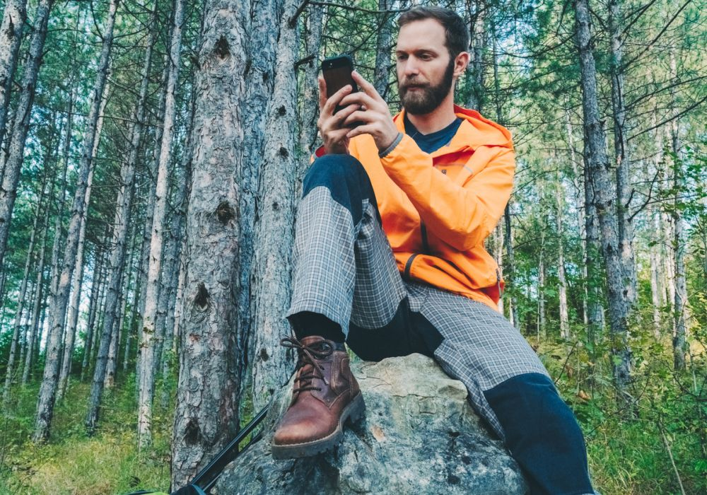 Stern looking man in an orange jacket sitting on a rock in a forest sending a text.  