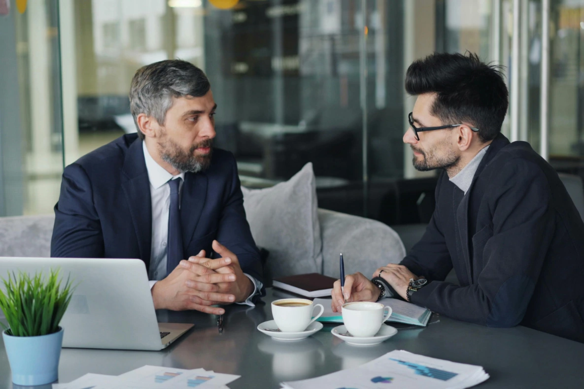 Two individuals in suits are having a discussion at a table with a laptop, coffee cups, and papers.