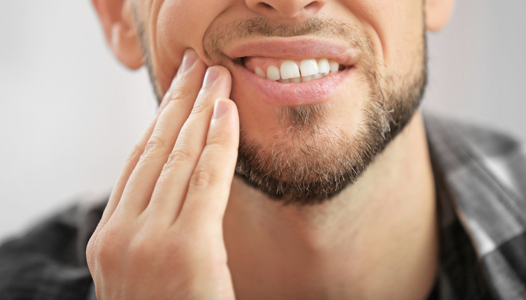 Mann med skjegg som holder seg til kinnet og viser ubehag, trolig på grunn av tannpine eller kjeveproblemer.