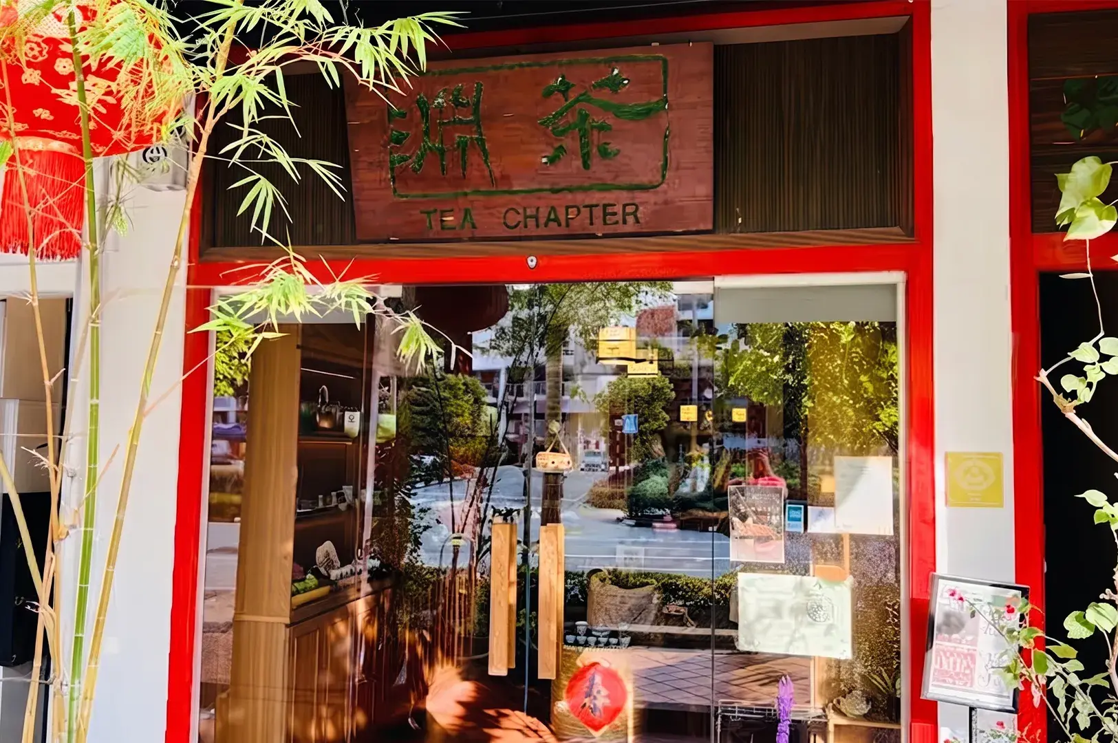 Exterior view of the "Tea Chapter" storefront featuring a wooden signage board, red door trim, glass entrance doors, and bamboo foliage.
