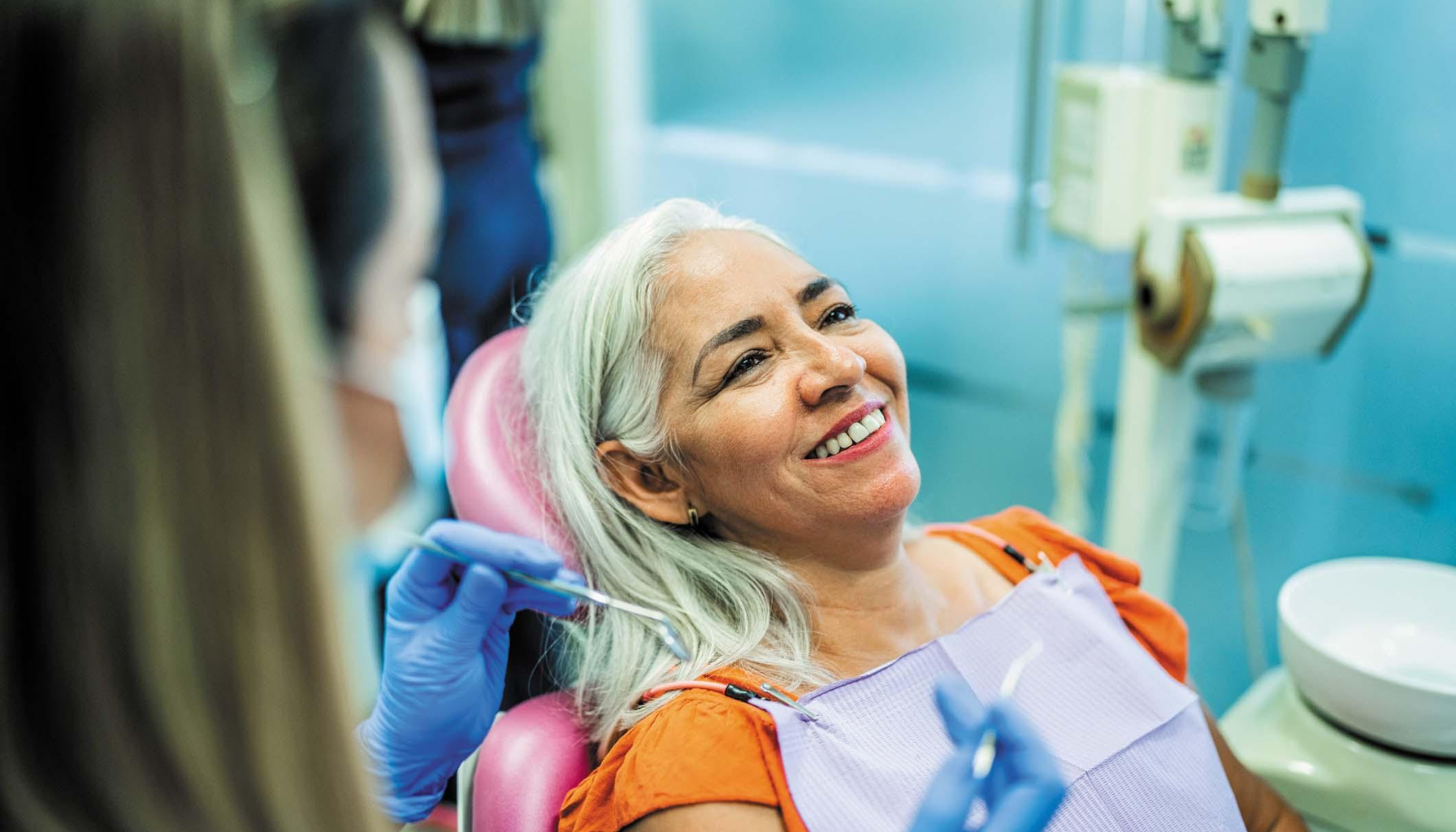 Eldre kvinne smiler i tannlegestol mens tannpleier forbereder undersøkelse med hansker og tanninstrumenter i klinikkmiljø.