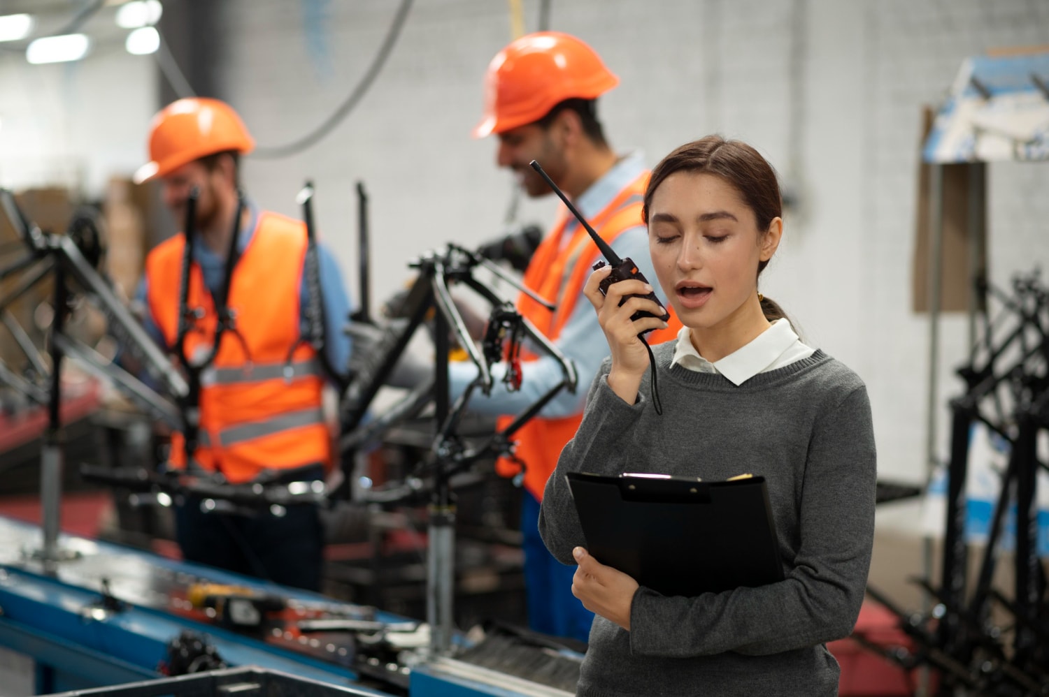 A woman holding a clipboard speaks into a walkie-talkie on a bicycle assembly line, with workers in safety vests behind her. Using UCaaS, she benefits from unified communications and cloud-based communication tools that connect frontline teams through cloud telephony and real-time updates.