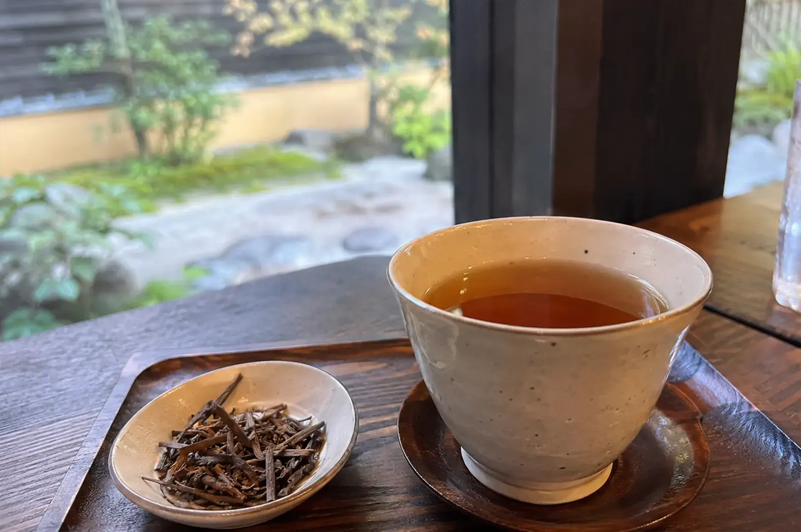 A warm cup of dark brown tea is served on a wooden tray alongside a small saucer containing a pile of dry, roasted tea stalks, with a blurry Japanese garden visible through the window in the background.