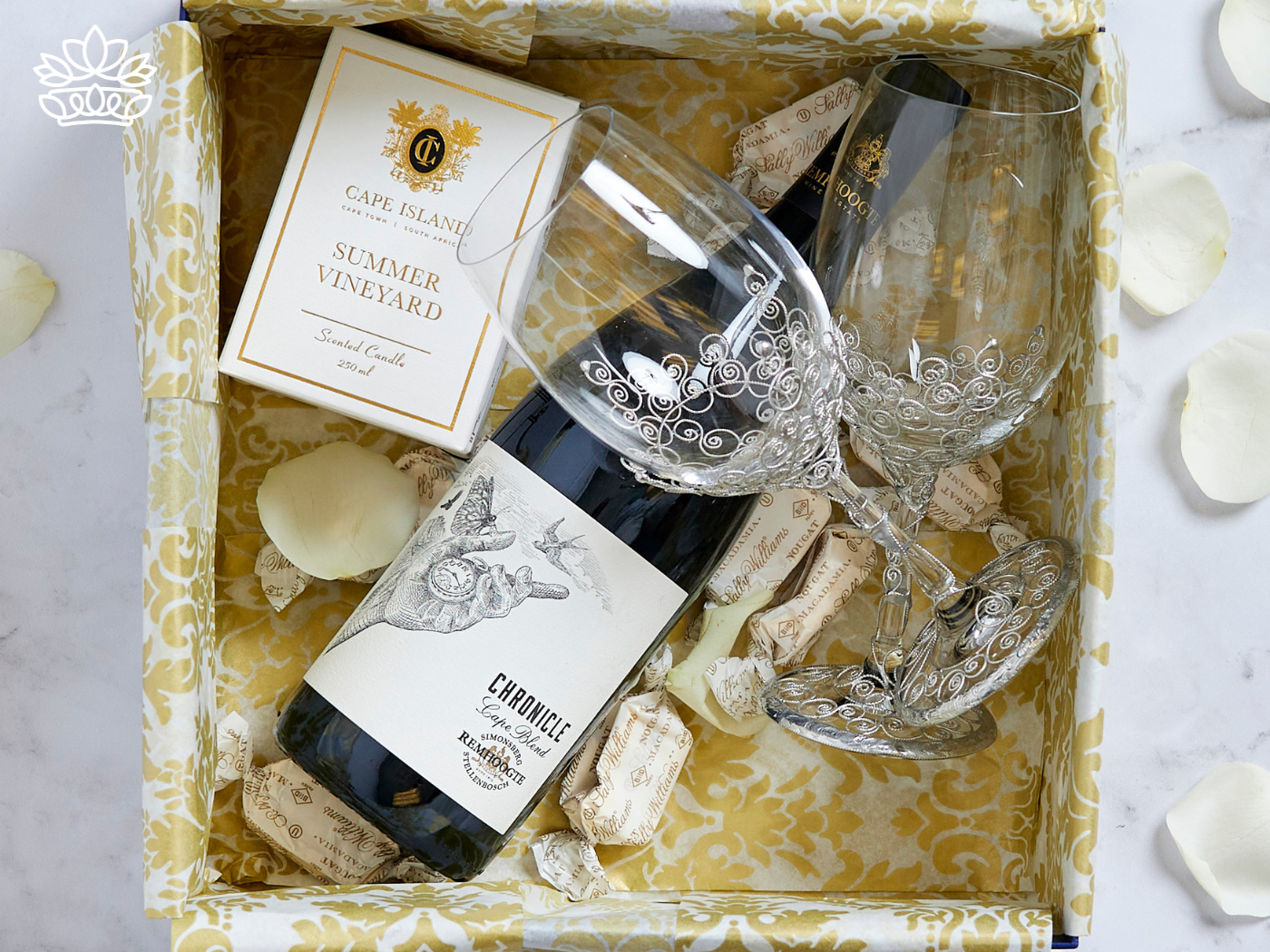 A beautifully arranged gift box containing a bottle of wine, two intricately designed wine glasses, a scented candle, and chocolates, set on patterned tissue paper with scattered rose petals. Fabulous Flowers and Gifts. Gift Boxes for Boyfriend. Delivered with Heart.