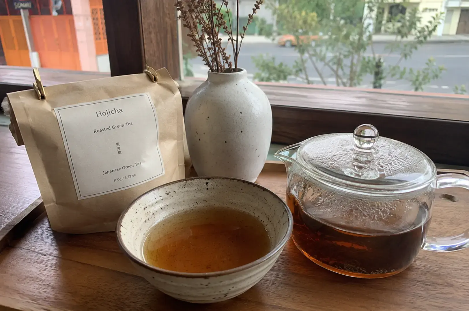 A light-brown paper package labeled "Hojicha" sits on a wooden tray alongside a glass teapot filled with amber-colored tea, a small ceramic bowl containing a warm infusion, and a decorative white vase holding dried sprigs, all placed by a sunlit window overlooking a street.