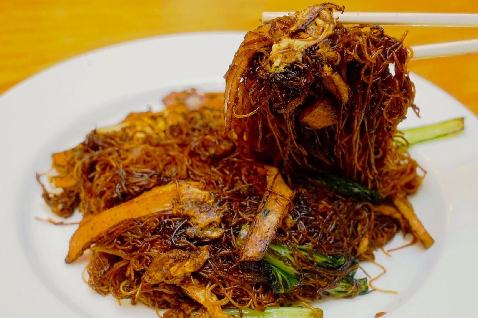 Stir-fried thin noodles with dark sauce on a white plate, lifted with chopsticks, accompanied by vegetables and sliced meat, create a savory allure.