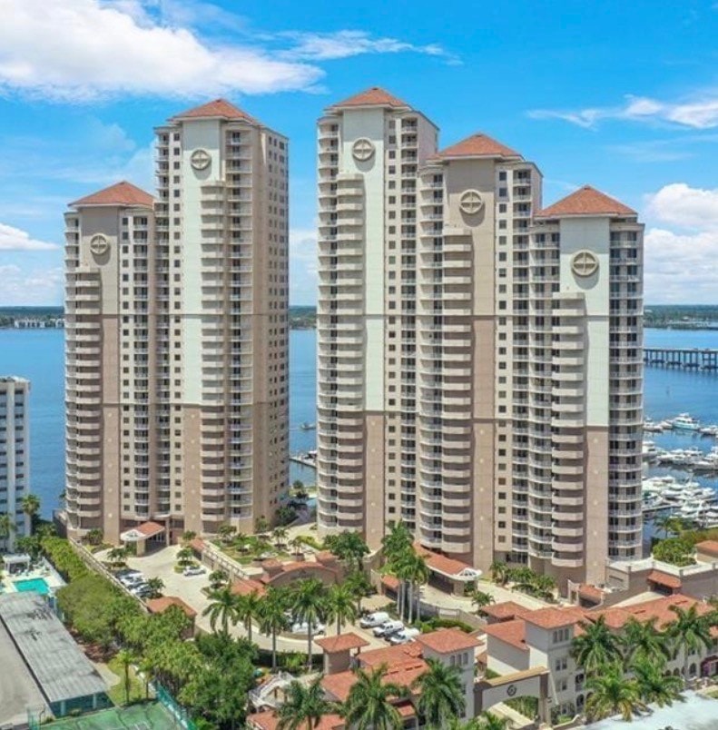 The image showcases a spacious luxury condo interior featuring floor-to-ceiling windows that offer breathtaking views of the Caloosahatchee River, set in a prestigious high rise at High Point Place in downtown Fort Myers. The neutral interior design includes granite countertops and crown molding, enhancing the luxurious lifestyle in this urban waterfront setting.