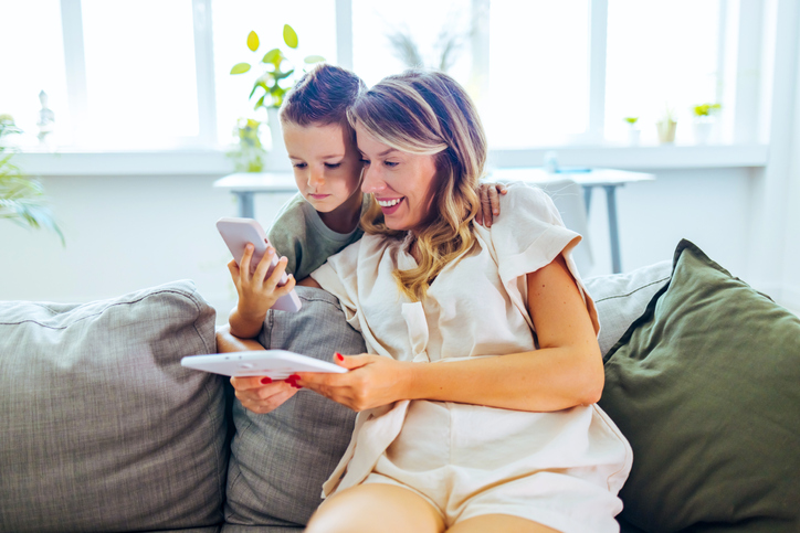 Cheerful mom and son sitting on a sofa looking at a cell phone and a tablet.