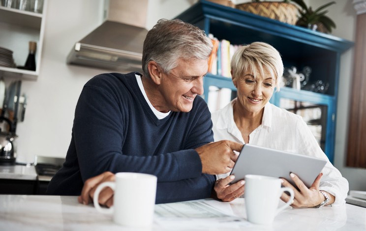 Handsome gray-haired man and blonde woman with short hair having coffee and looking at a tablet.