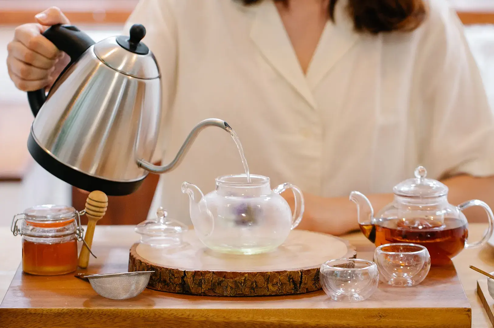 Person pouring hot water from a stainless steel gooseneck kettle into a clear glass teapot during a tea preparation ritual.