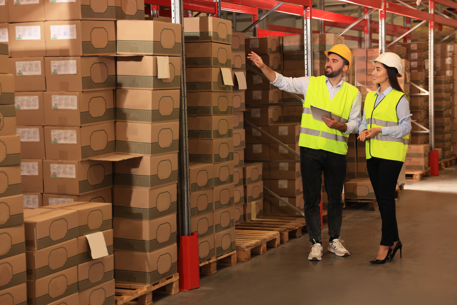 Two people wearing hard hats and high-visibility vests stand in a warehouse aisle, surrounded by stacks of cardboard boxes. One person holds a tablet and gestures towards the boxes.