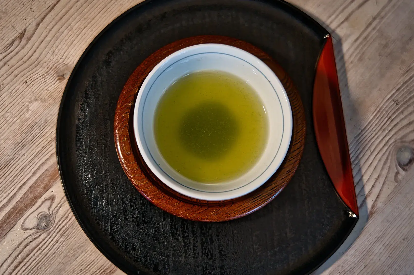 Top-down view of a small ceramic bowl of pale green tea served on a wooden saucer and a dark black tray.