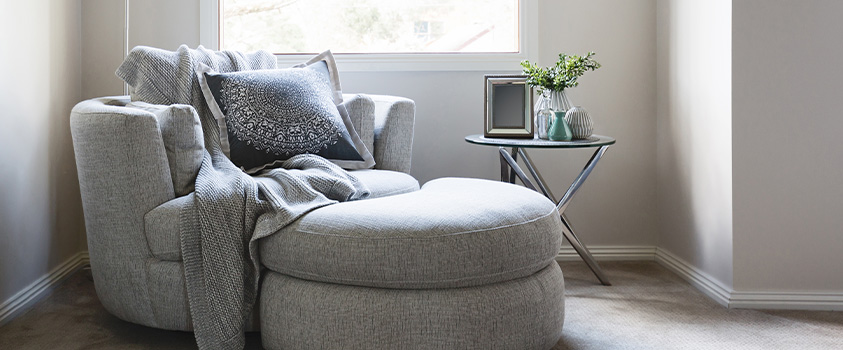 A circular shaped armchair and ottoman set with a grey blanket, white mandala cushion, and a glass and metal coffee table. Set in a bright corner space.