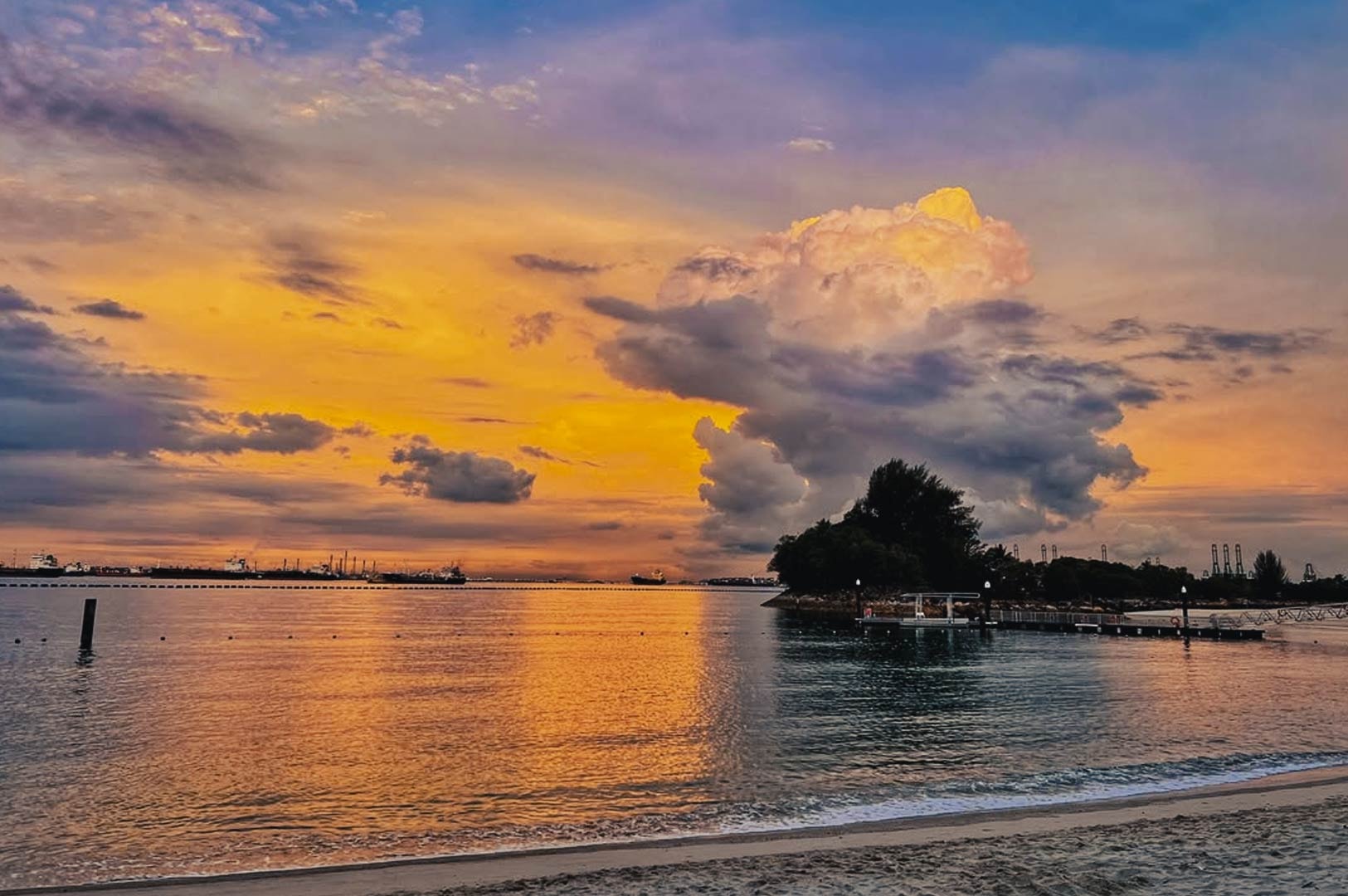 Colorful sunrise sky over Sentosa beach with calm waves and a small jetty in the distance.