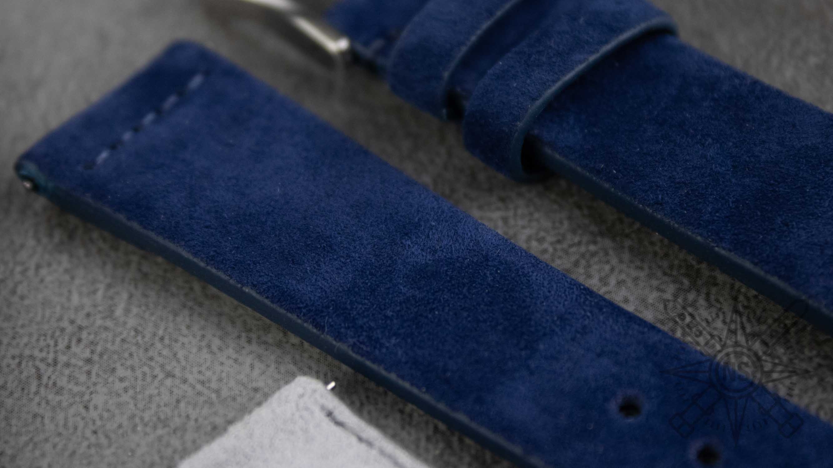 The image features an assortment of buckles in various finishes, including steel, gold plated, and black PVD, displayed alongside high-quality blue suede watch bands. This collection of suede straps showcases both elegance and durability, perfect for enhancing any timepiece.