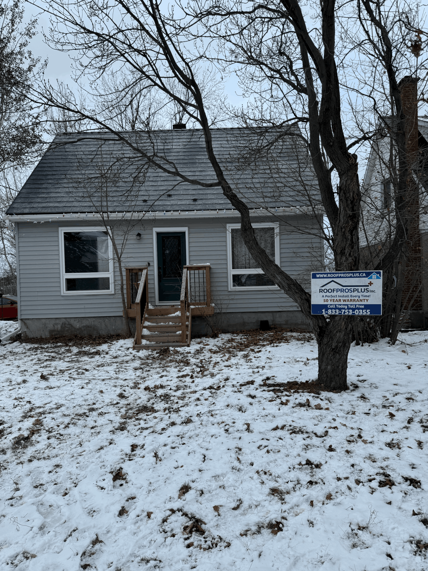 A residential house with a grey metal roof, featuring a Roof Pros Plus sign on a tree in the front yard.