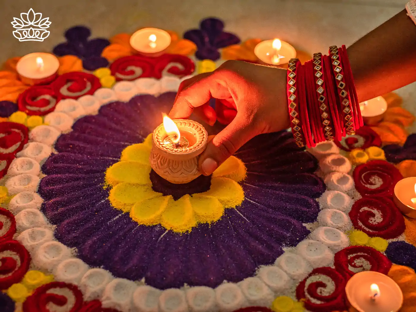 A person lighting a traditional clay Diwali lamp on a vibrant rangoli decorated with colorful flower patterns and candles, symbolising the festival of lights. Fabulous Flowers and Gifts - Diwali Flowers. Hindus celebrate this sepcial day.