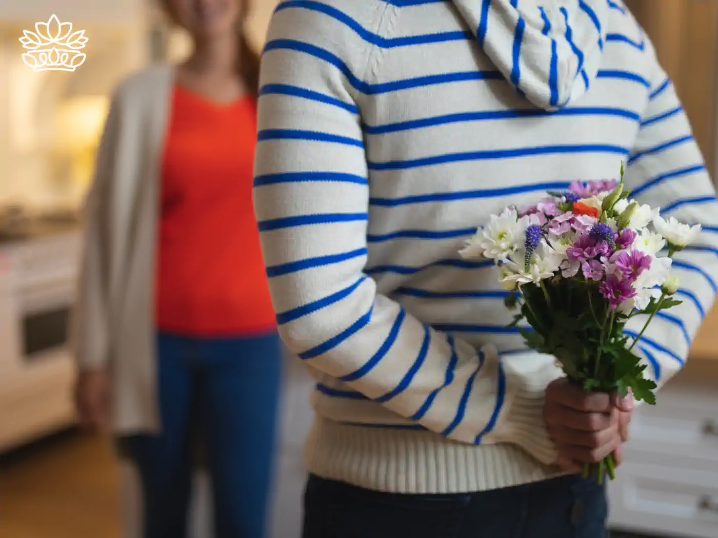Man in a striped sweater holding a bouquet of fresh mixed flowers, including white, purple, and pink blooms, with a smiling woman in the background in a cozy home setting. 