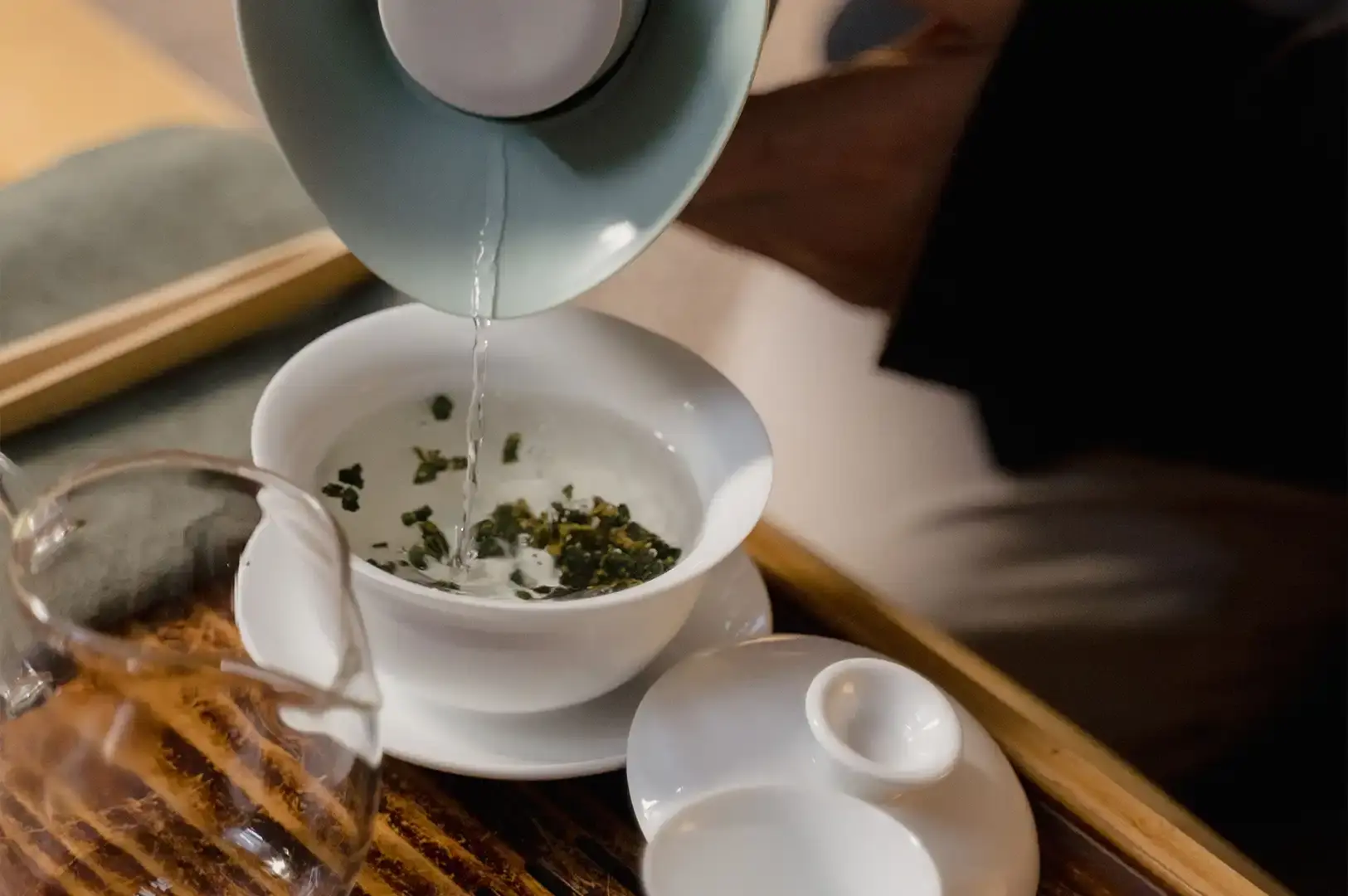 In this close-up, a light green teapot is tilted, pouring a stream of steaming hot water into a white ceramic cup that already contains loose Tie Guan Yin tea leaves. The scene is set on a wooden table, with other tea-making accessories visible, capturing the precise and ritualistic process of brewing this traditional Chinese Oolong tea.