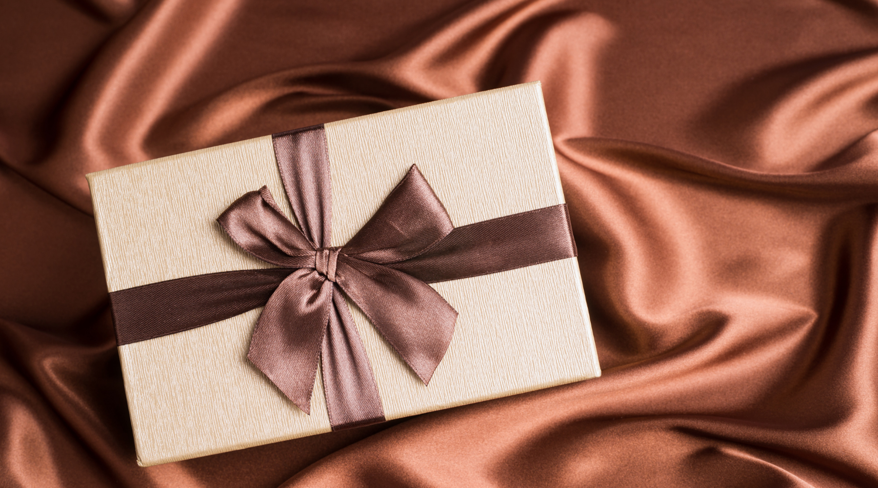 Gift box with beige texture and chocolate satin ribbon on a silky bronze fabric background.