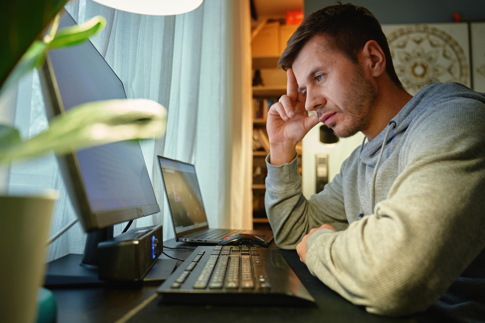 Man in a gray hoodie sitting at a home workstation, looking focused at a desktop screen with hand on forehead, representing remote work challenges and the need for reliable business phone systems.