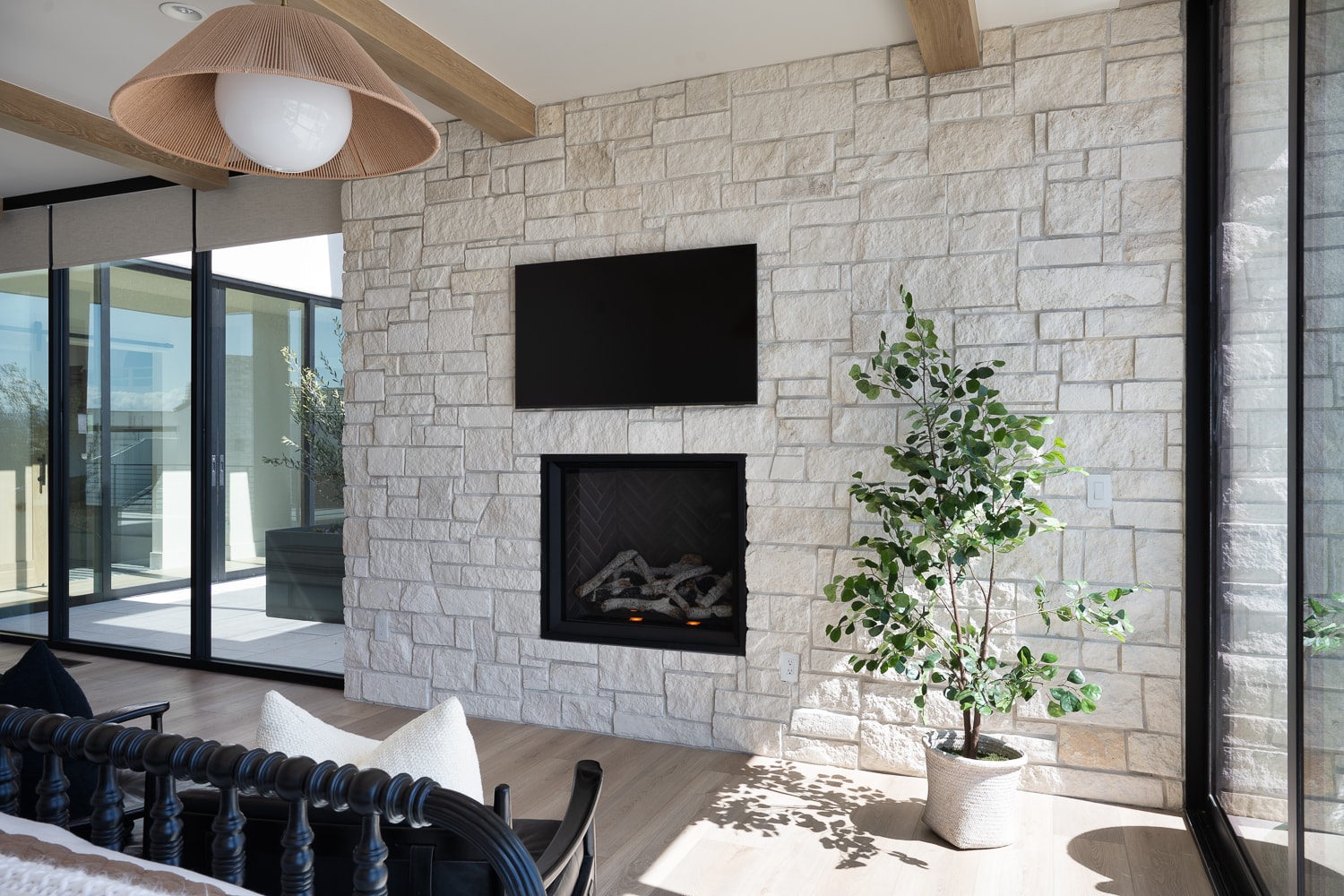 Minimalist living space featuring a woven pendant light, stone fireplace, modern black-framed windows, and a potted green plant on light hardwood floors.